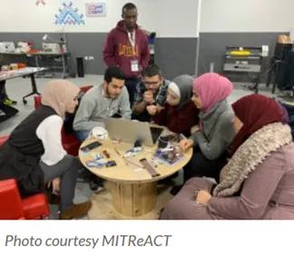 A group of women and men are sitting on a table, looking at a computer. One man is standing behind the group. The setting seems to be a classroom.