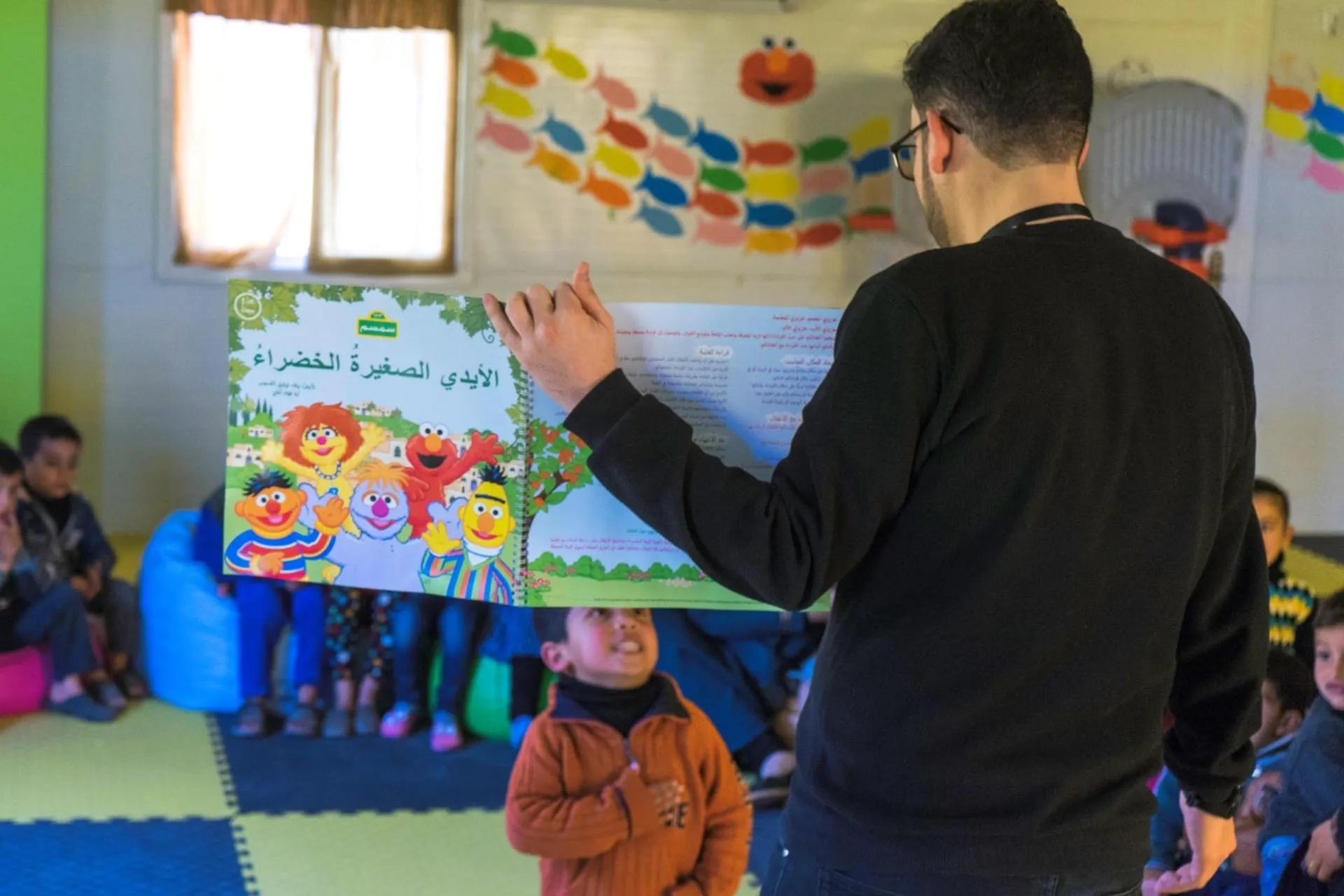 A man shown from behind is showing a book to young children in a classroom