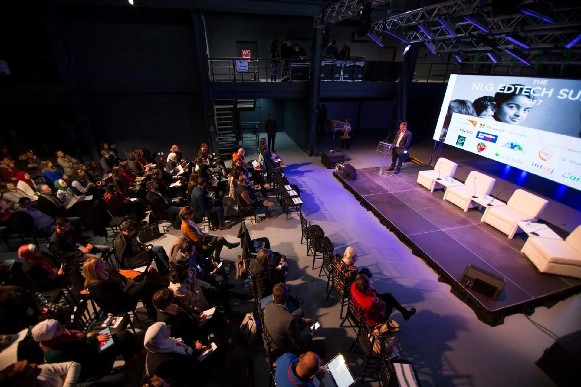 A man is talking in front of a crowd in an amphitheater, with the NLG EdTech PowerPoint behind him