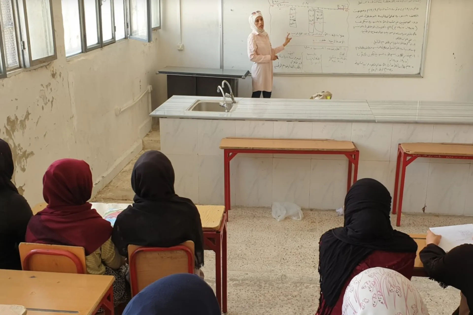 The picture shows a female teachers standing by a white board teaching a group of adolescent girls.