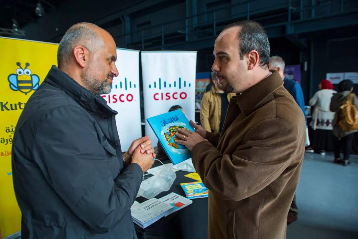 Two men discussing in a front of a booth, with one of them holding a book untitled "I am human" (in arabic)