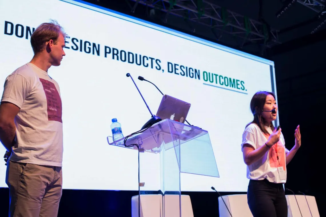 A woman and a man presenting a powerpoint that says "Don't design products, design outcomes"