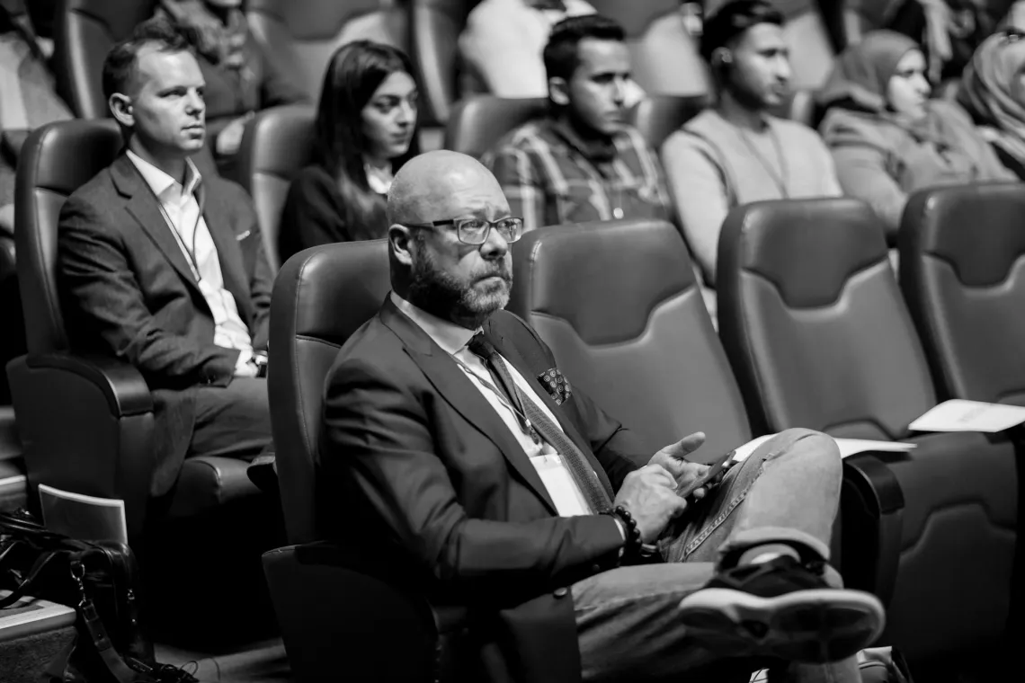 A black and white picture of a man sitting in the front row of an amphitheater, with other participants sitting behind him.