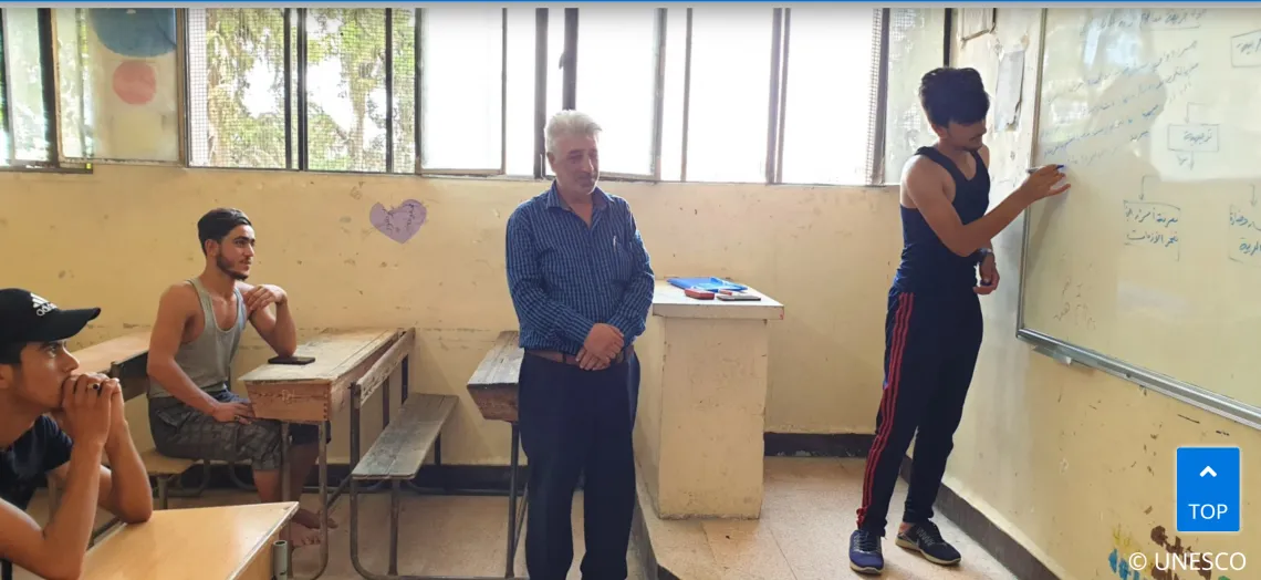 The picture shows an adolescent boy writing on a white board, his teacher standing up and other adolescent boys sitting behind him