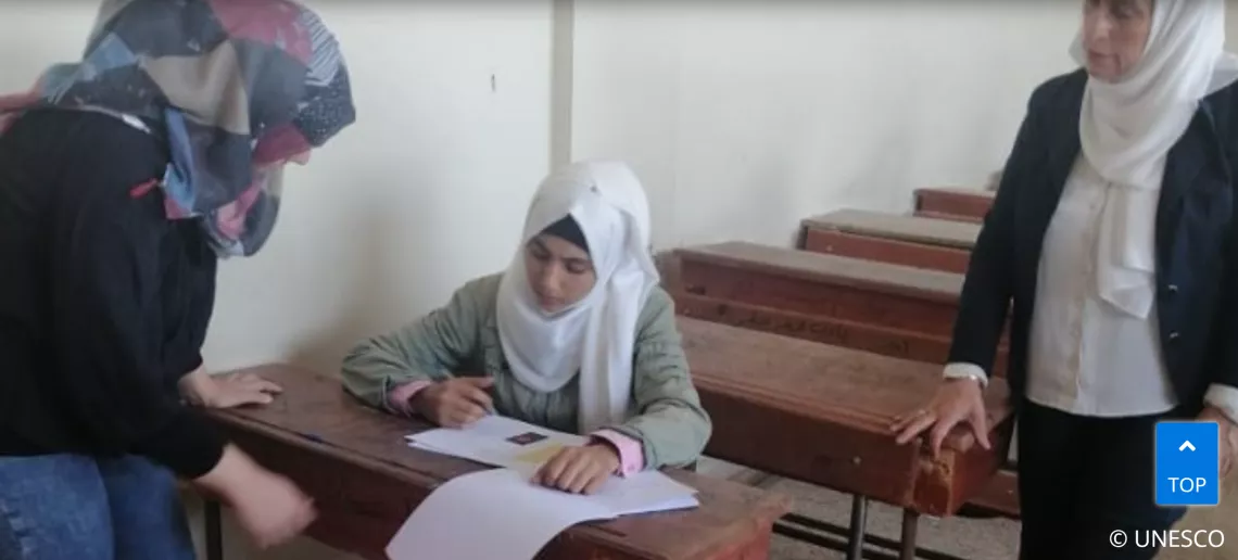 A young girl is writing on a paper while her two female teachers are standing besides her in a classroom