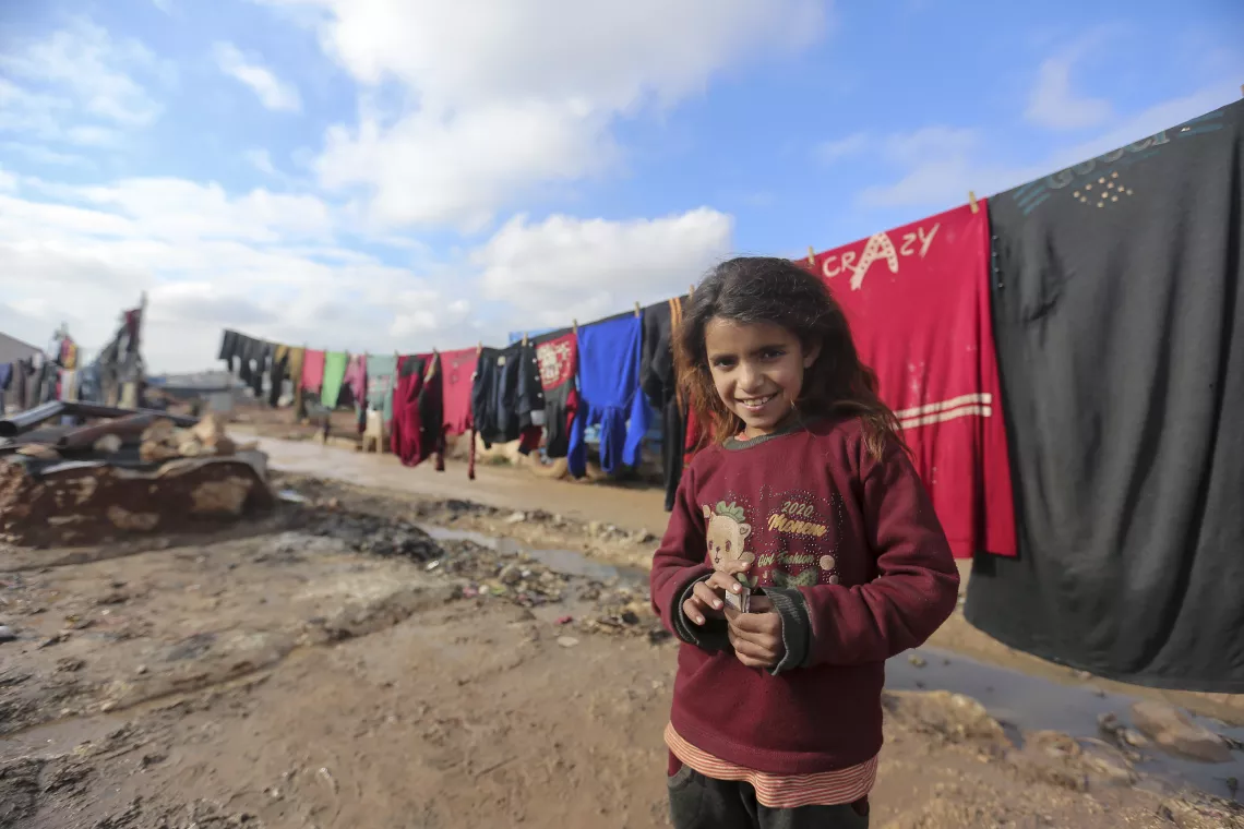 A girl is smiling and standing in front of a laundry line, outside in a refugee camp